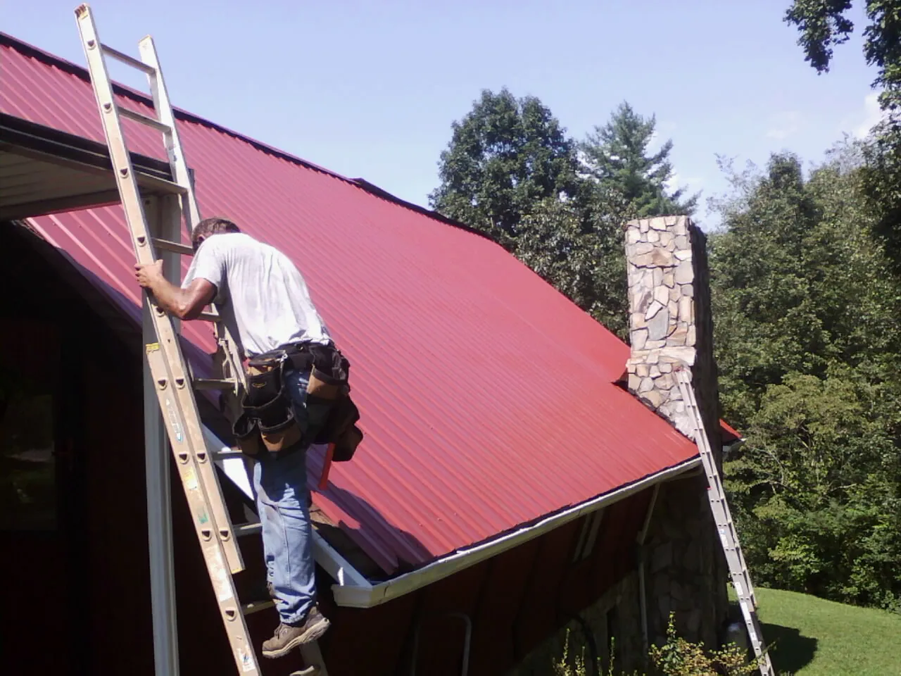 Roofer on a ladder beside a red metal roof and stone chimney — Soffit & Fascia Repair by Roofing Brothers in Whitesville